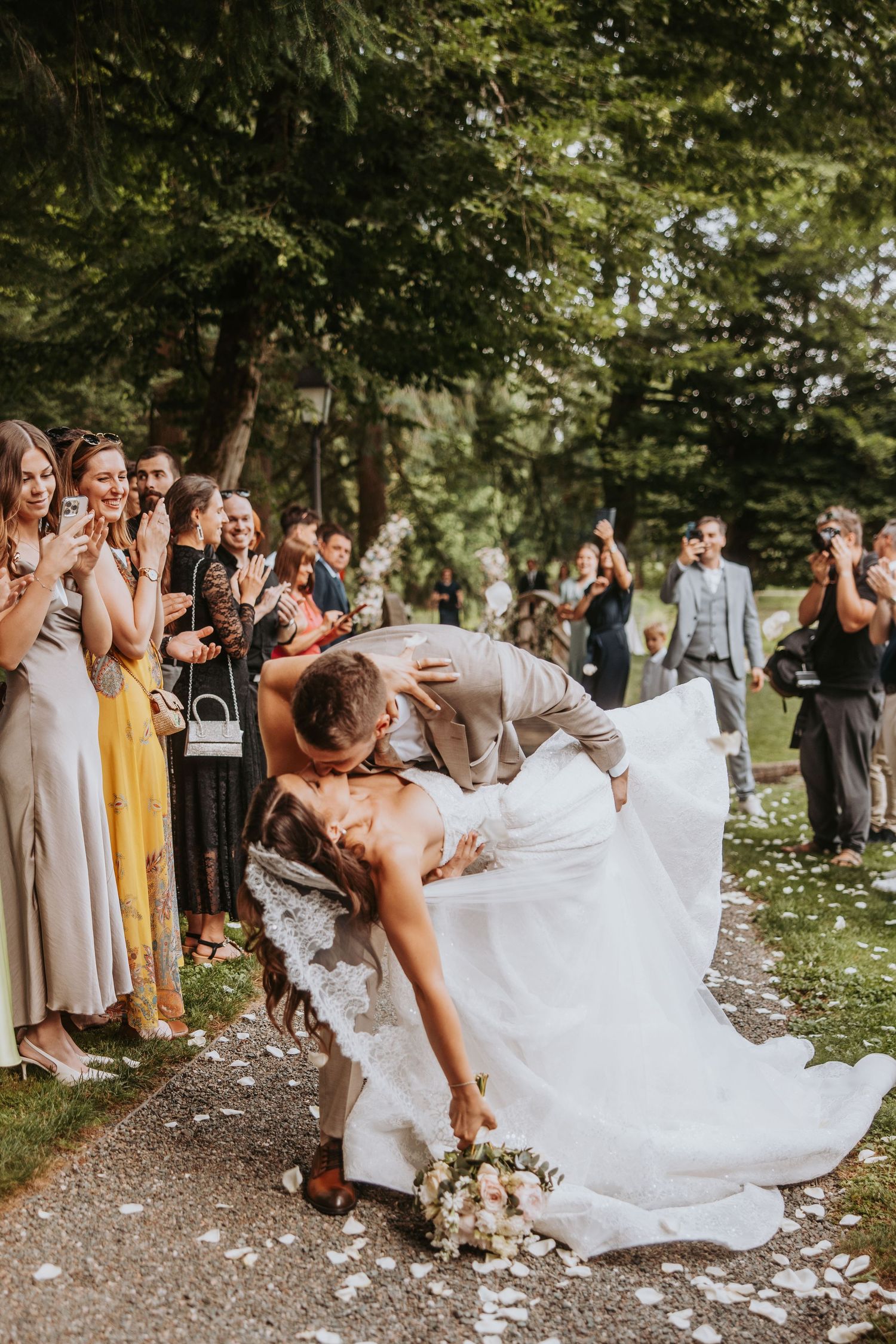 Groom dipping bride for romantic kiss outdoors while wedding guests cheer and celebrate.