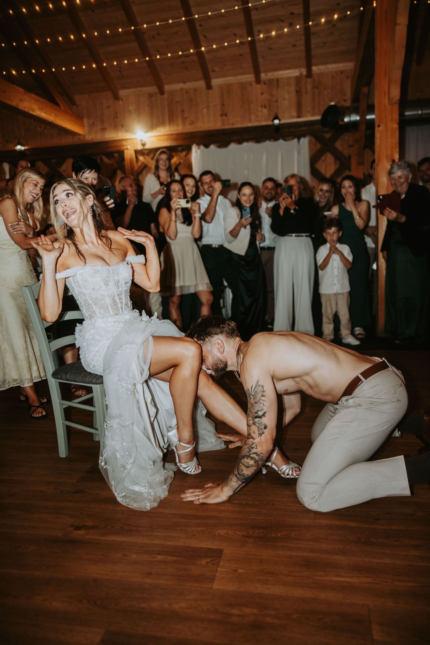 Groom removing garter from bride's leg while guests cheer and photograph at a rustic barn reception.