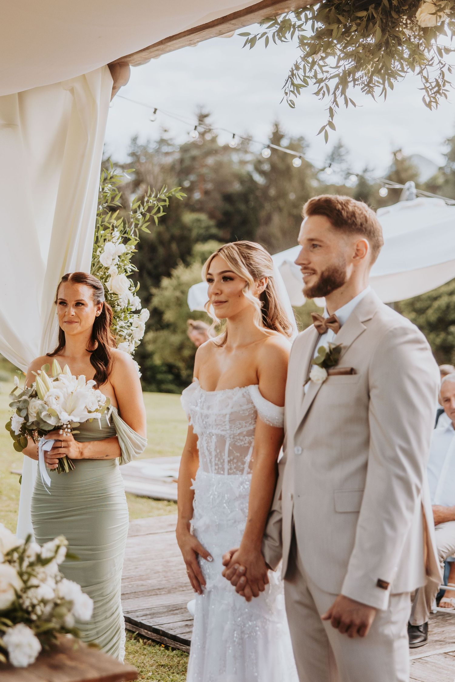 Bride and groom holding hands at outdoor wedding ceremony with bridesmaid in sage green.