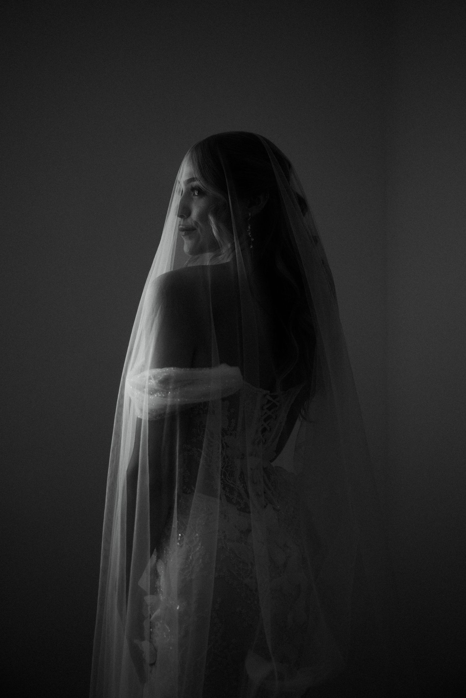 Elegant bride in a lace wedding dress draped with a sheer veil, captured in a dramatic black and white portrait.
