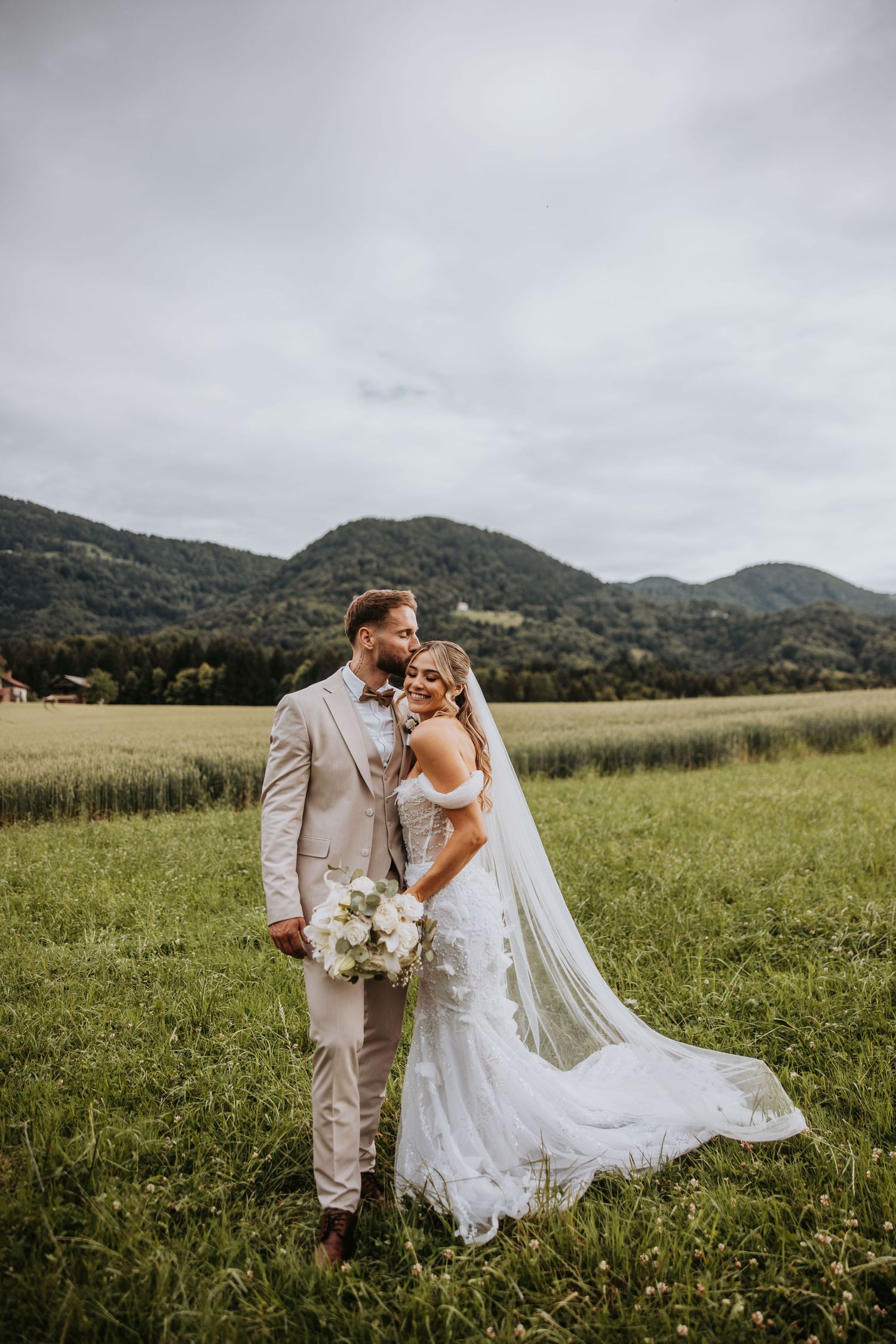 Bride and groom posing romantically in a green meadow with stunning mountain backdrop on their wedding day.