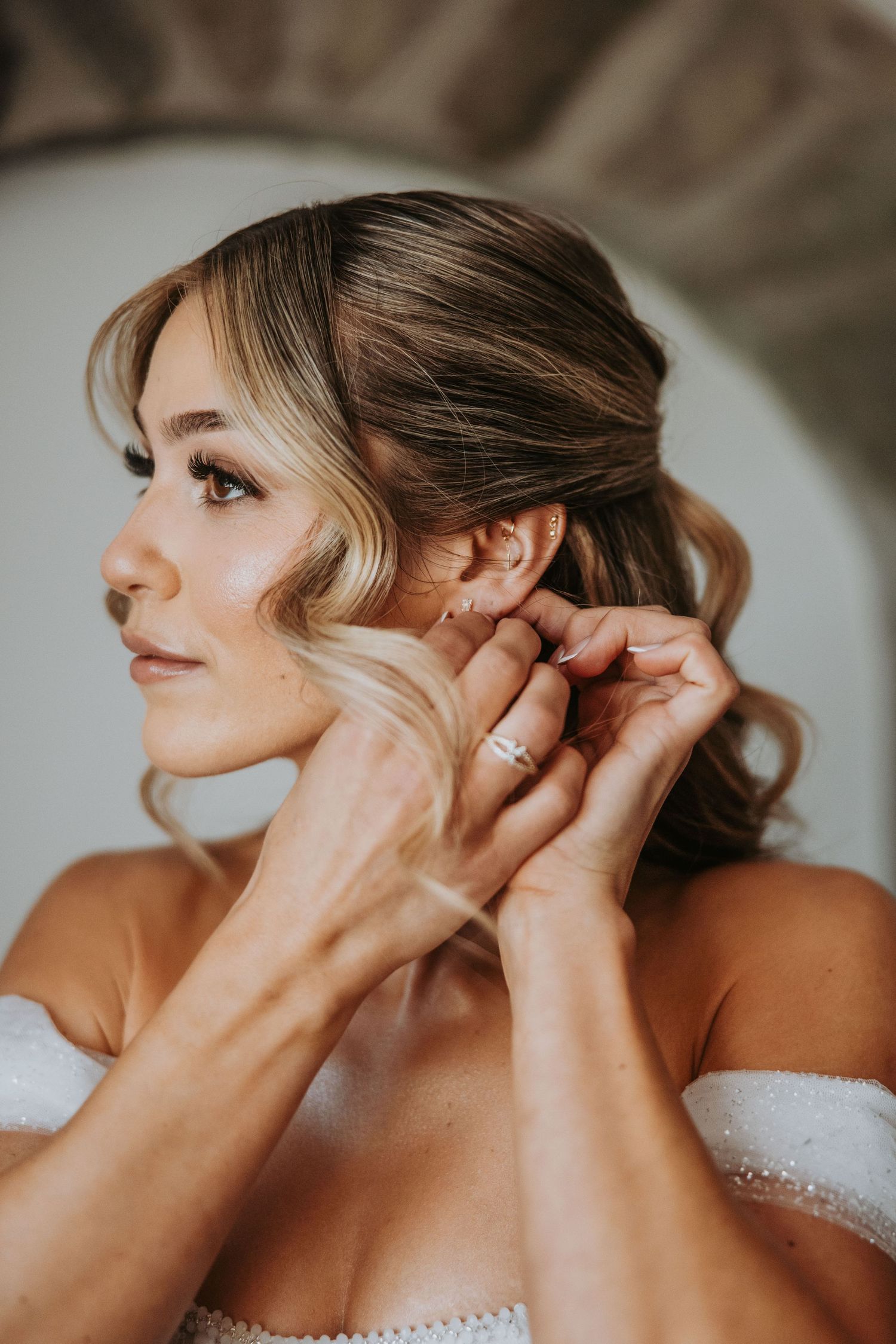 Close-up of bride having earring placed while wearing elegant updo hairstyle and off-shoulder wedding dress.