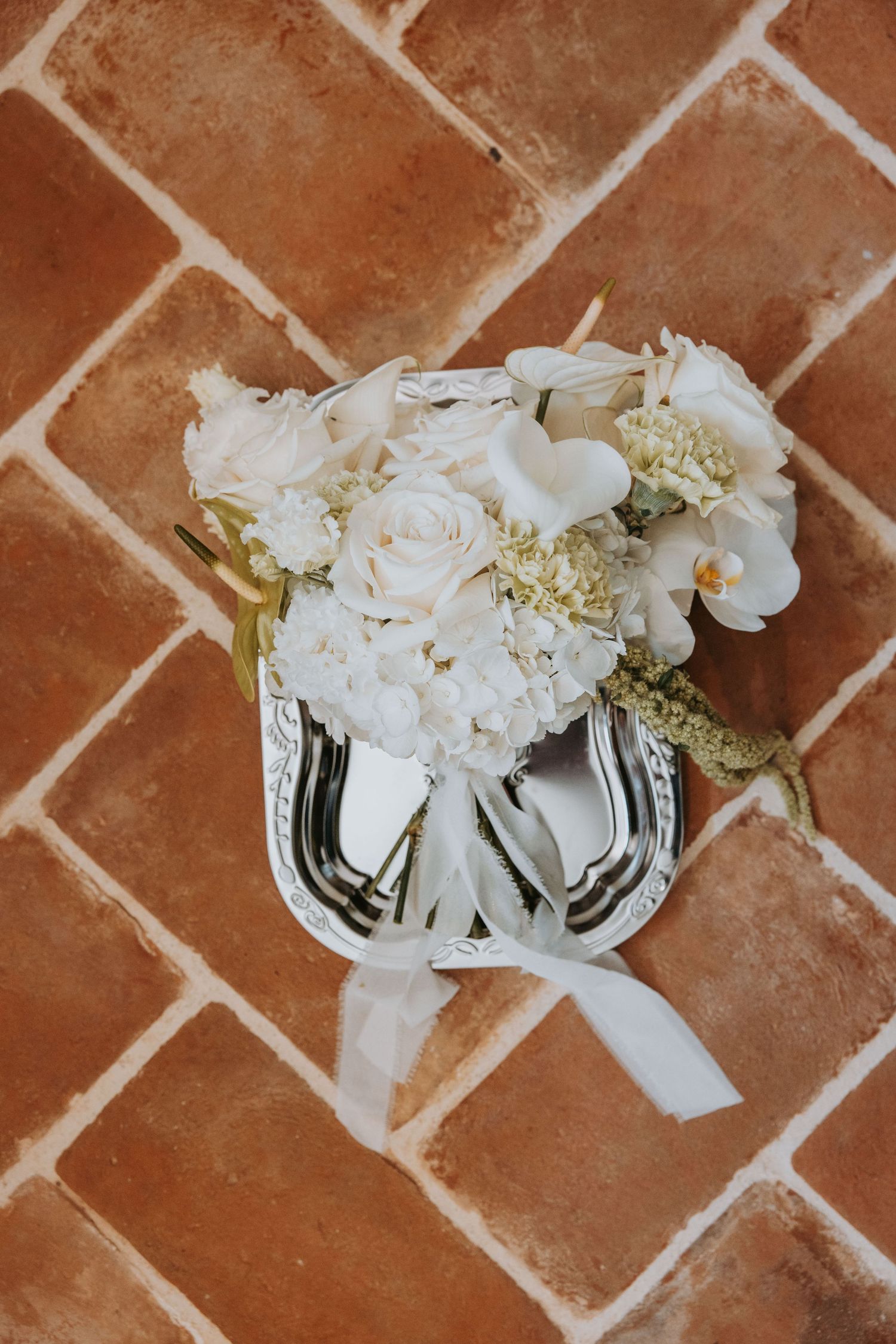 White wedding bouquet with roses, hydrangeas, and orchids in a silver vase on terracotta tile floor.
