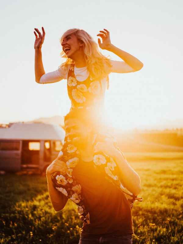 Joyful woman in a floral dress sitting on someone's shoulders in a golden sunset field.