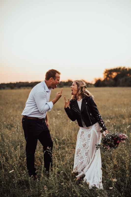 Bride in lace dress and leather jacket laughing with groom in a golden sunset meadow holding wildflowers.