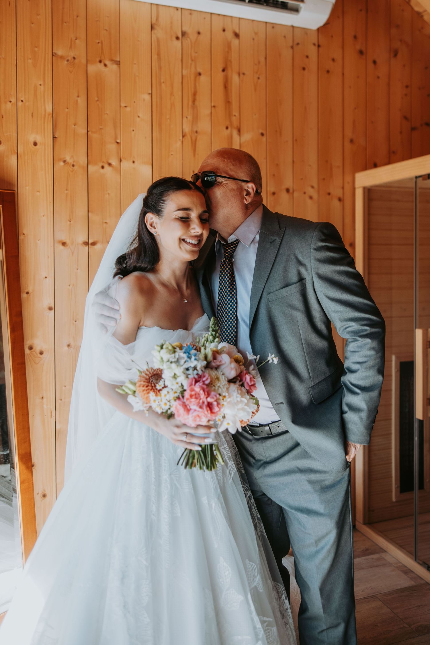 Bride in white gown holding colorful bouquet smiles as man in gray suit kisses her cheek in wood-paneled room.