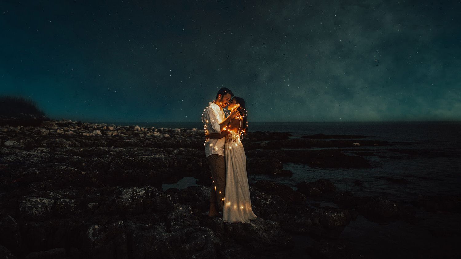 Wedding couple embracing under a starry night sky on a dramatic rocky coastal landscape.