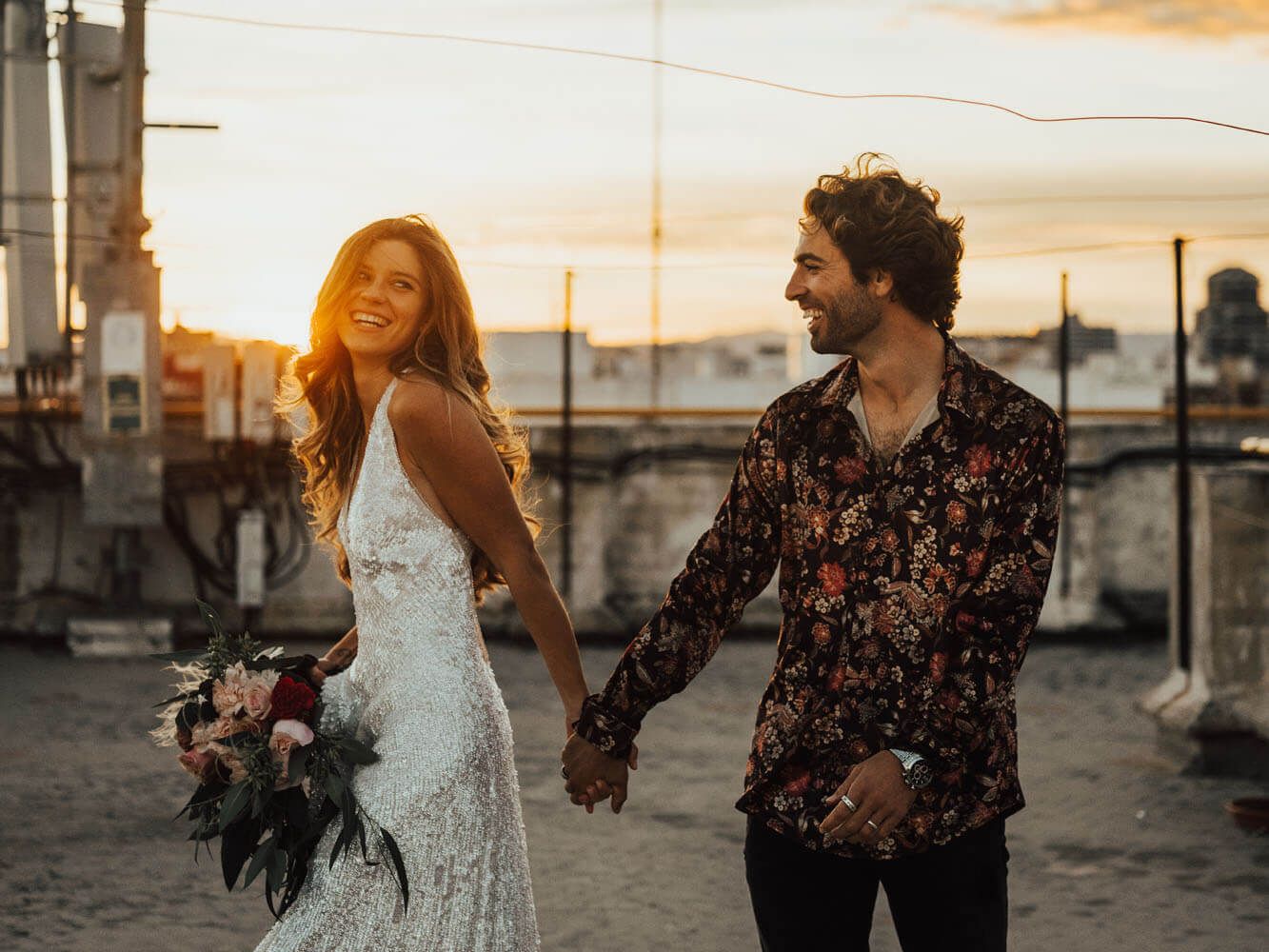 Smiling bride in lace gown and groom holding hands on rooftop at golden sunset with bridal bouquet.