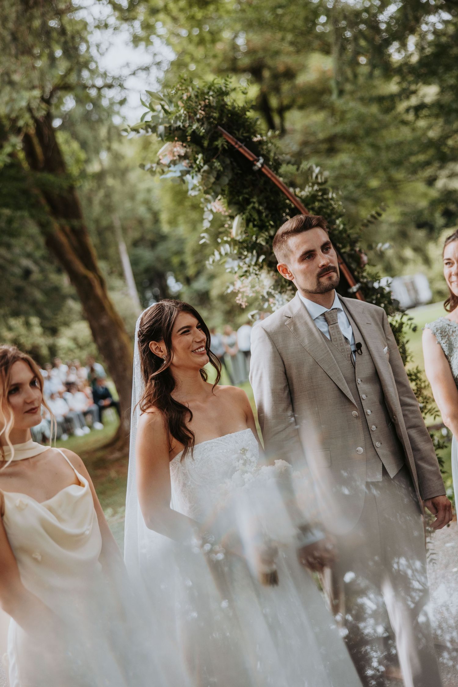 Smiling bride in a white gown holds hands with groom in a beige suit during an outdoor garden wedding ceremony.