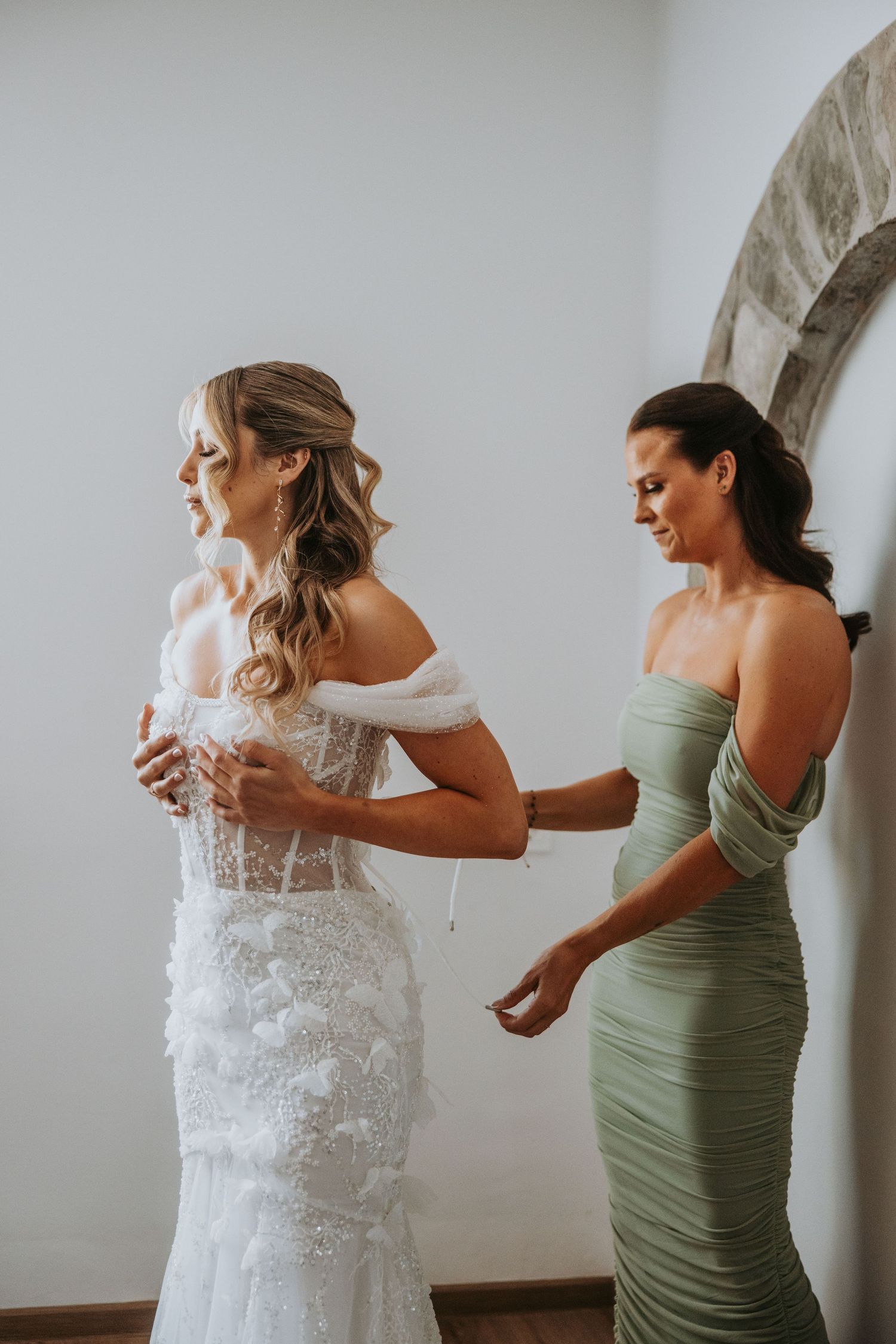 Bridesmaid in sage green gown helping bride button her lace off-shoulder wedding dress before ceremony.