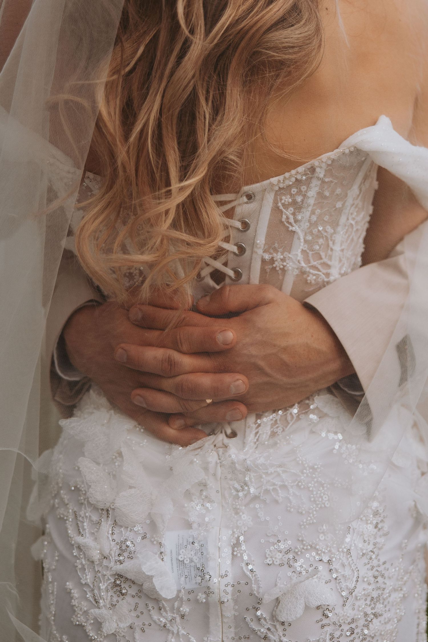 Close-up of groom's hands tenderly embracing bride's lace corset wedding dress from behind.