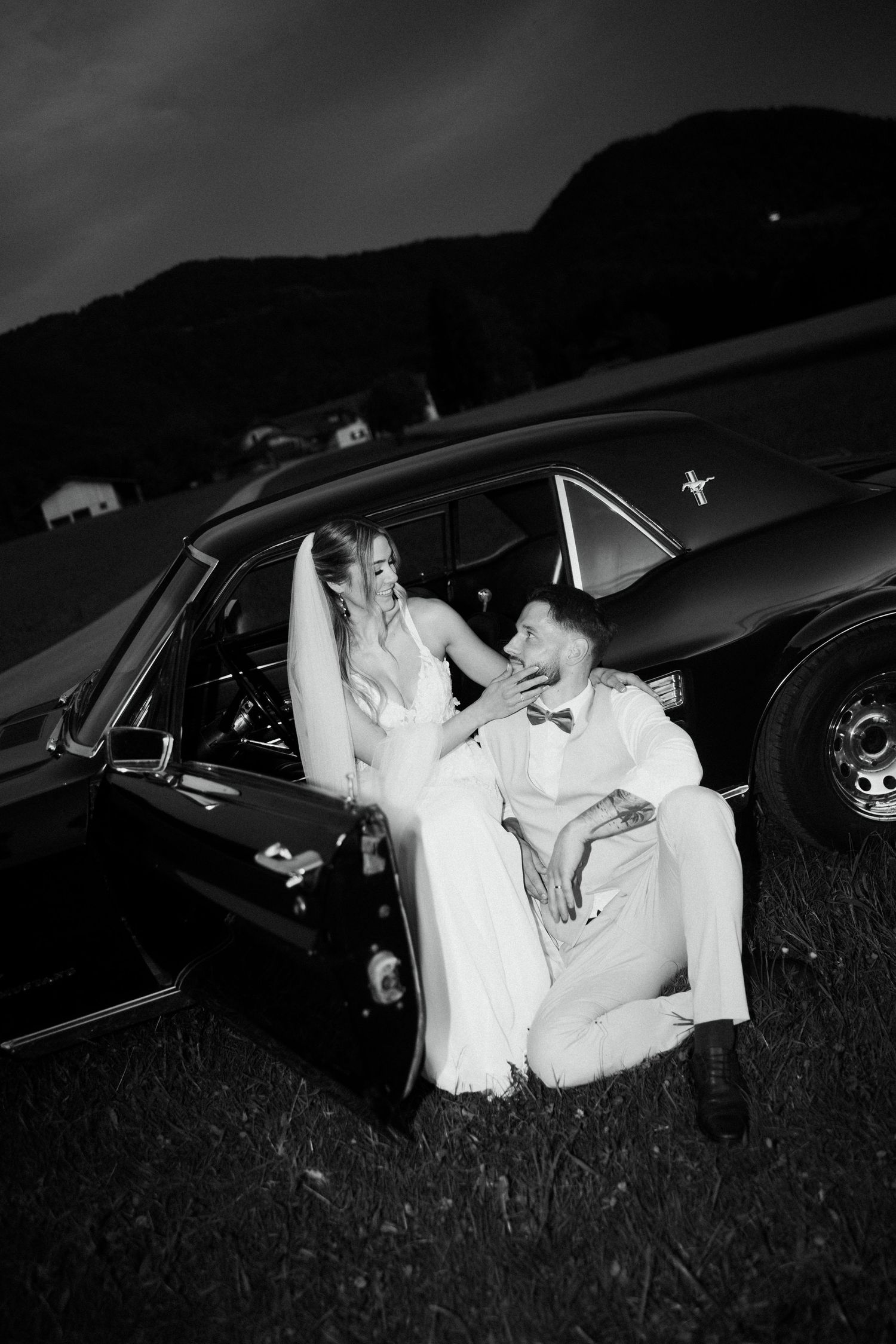 Black and white photo of bride and groom posing beside a classic Ford Mustang at night.