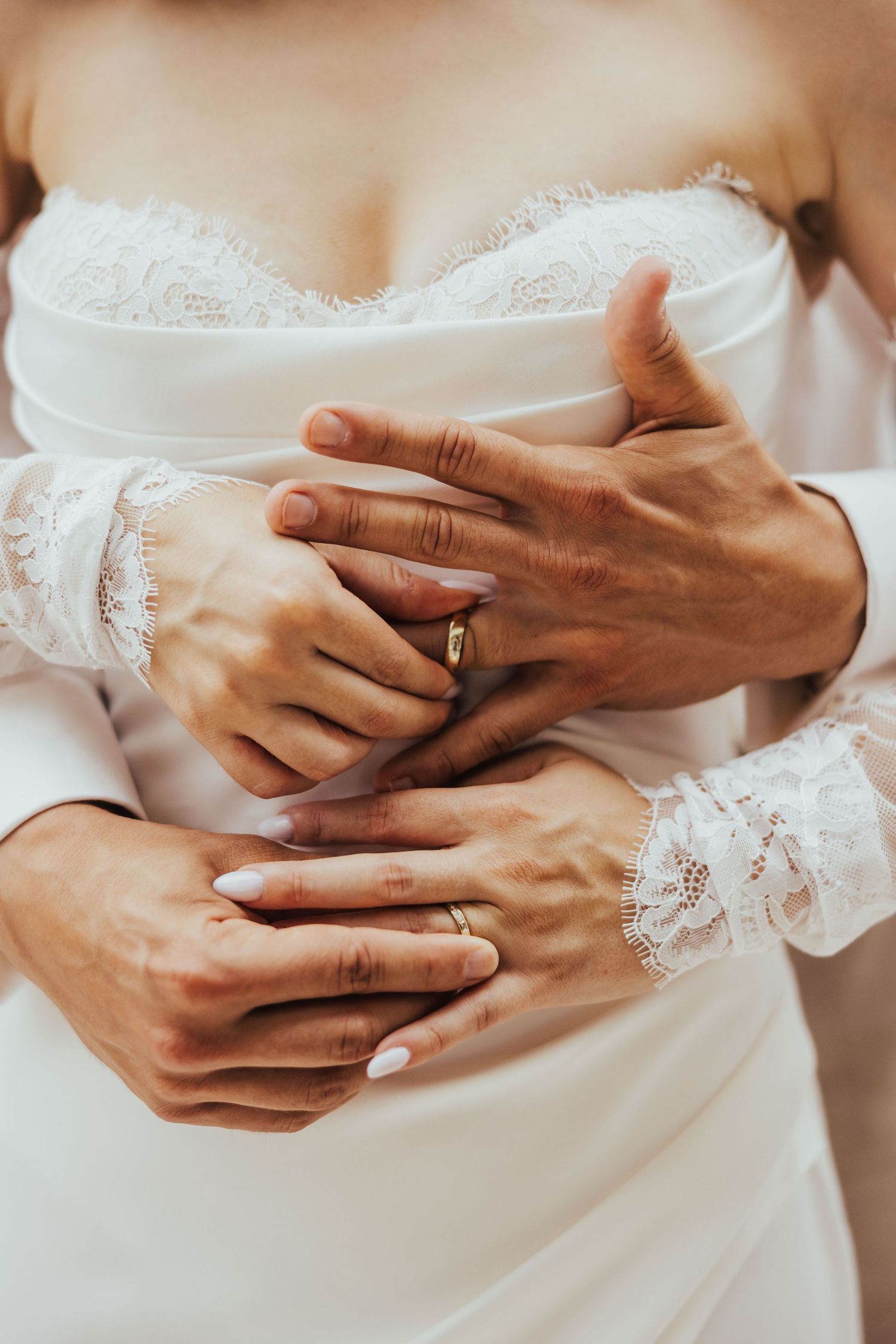 Close-up of bride and groom holding hands together showing their wedding rings, with lace wedding dress detail visible.