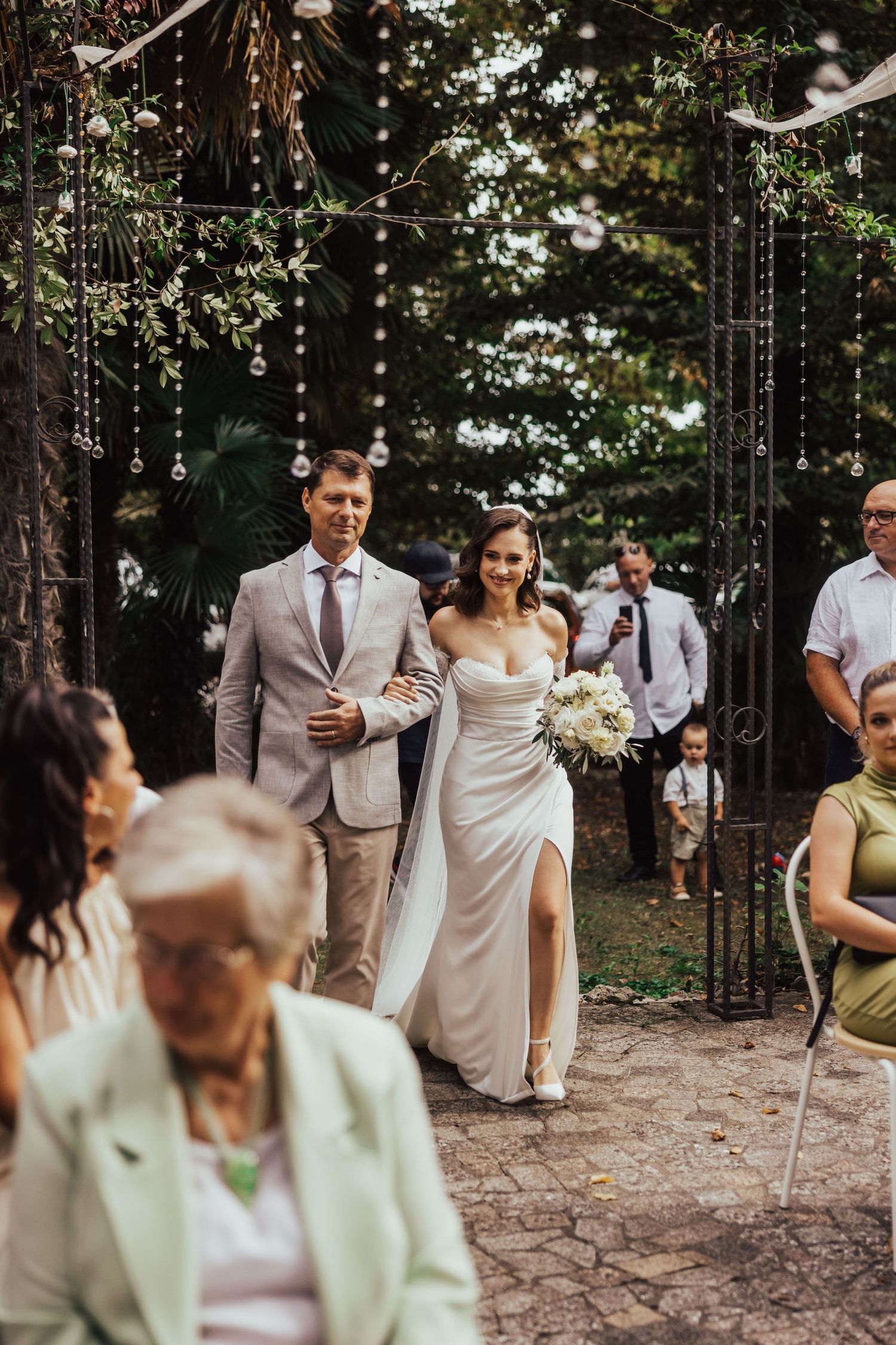 Bride in white gown holding bouquet walks down outdoor aisle with escort at garden wedding.