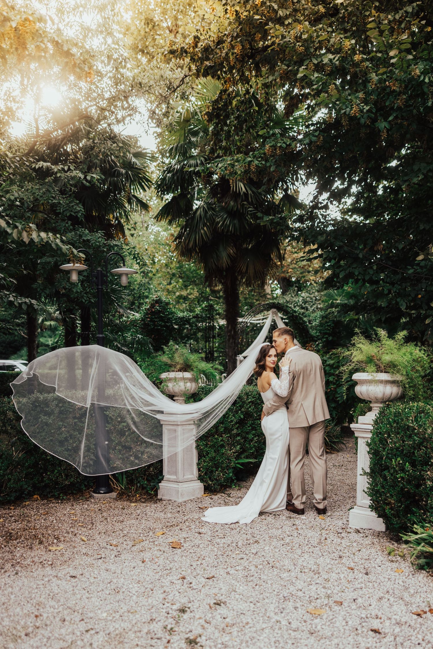Bride and groom embrace in lush garden with flowing veil, surrounded by elegant stone urns and tall trees in golden light.