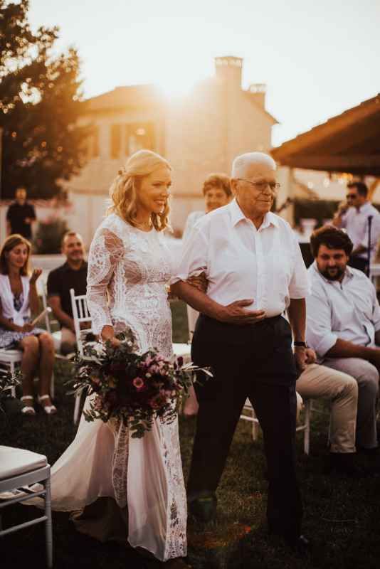 Bride in lace gown walks down outdoor aisle with elderly gentleman at golden hour wedding ceremony.