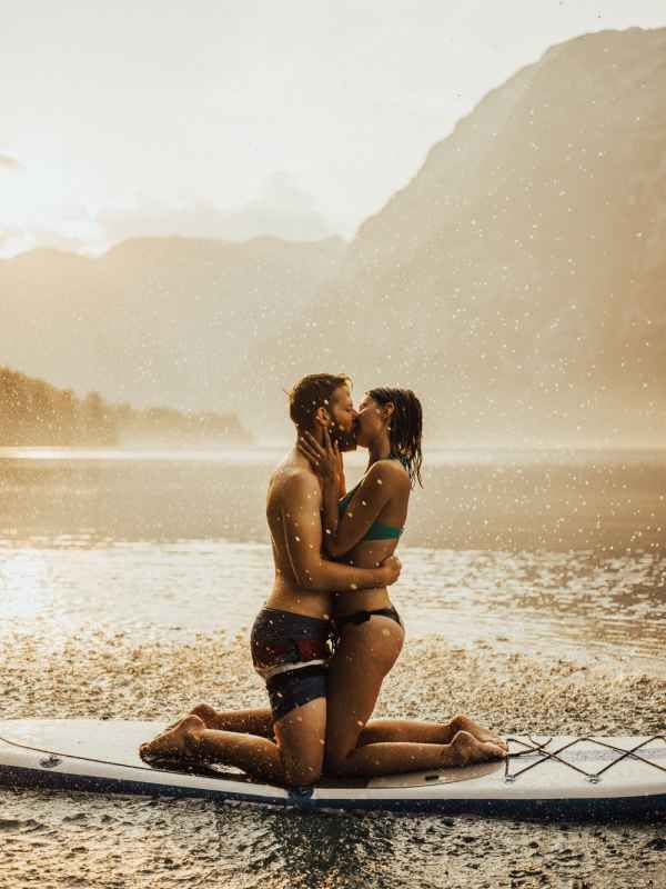 Couple sharing passionate kiss on paddleboard at golden sunset lake with misty mountain backdrop.
