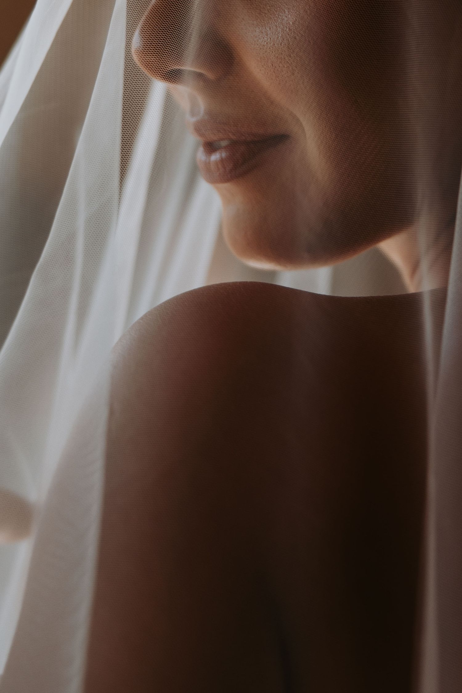 A bride wearing a white veil draped over her bare shoulder, captured in soft warm light.