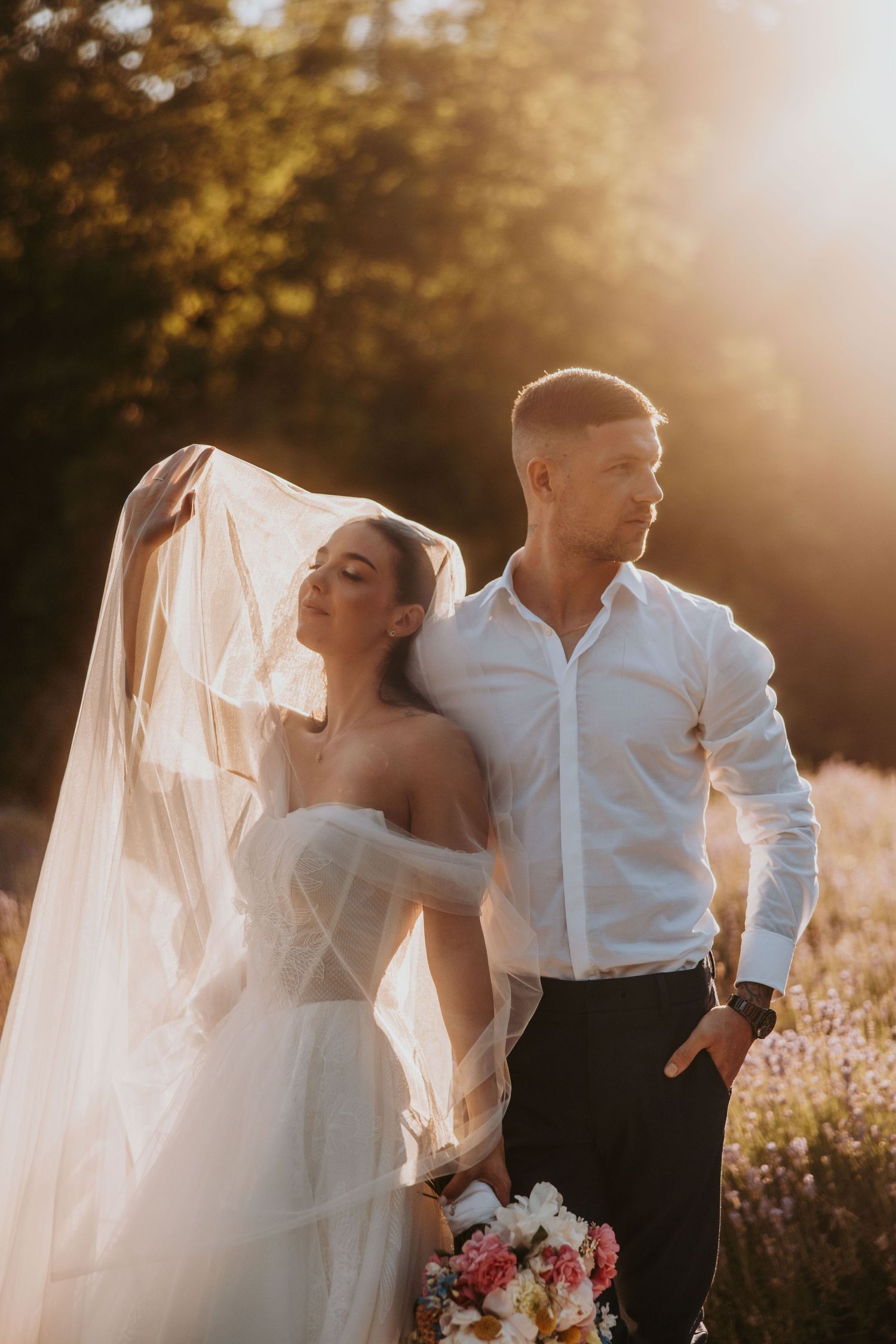 Bride and groom standing in golden sunset light in lavender field, veil flowing in the breeze.