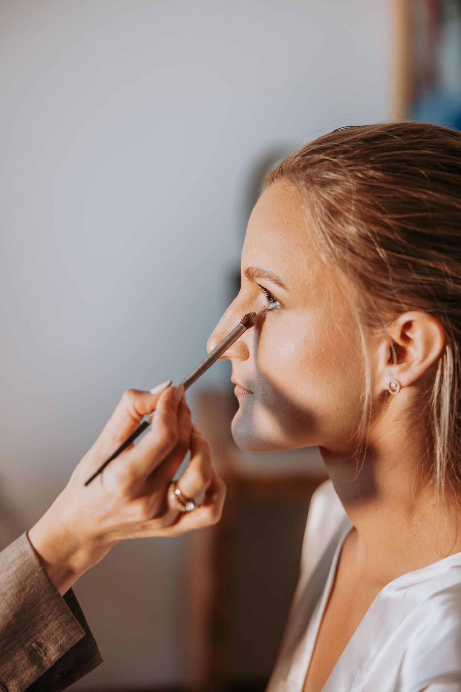 Professional makeup artist applying eyeshadow to a blonde bride during wedding morning preparations in natural light.
