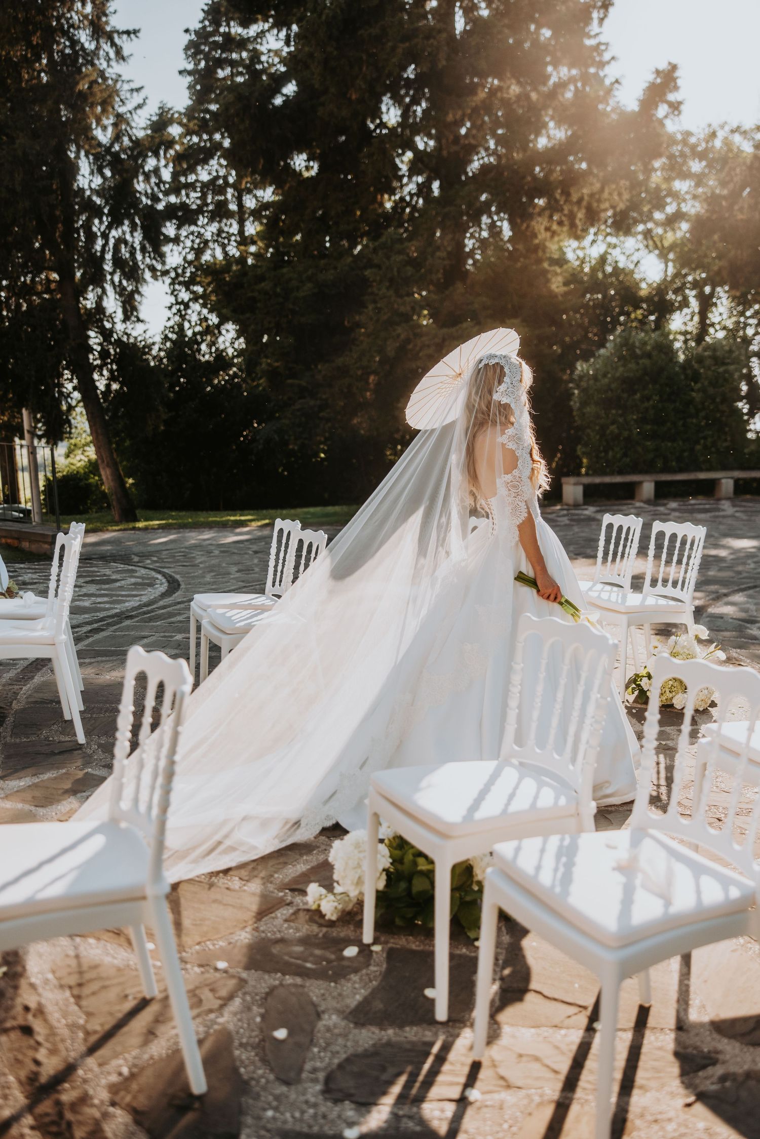 Bride in a flowing white gown and veil walks through an outdoor wedding venue with white chairs in sunlight.