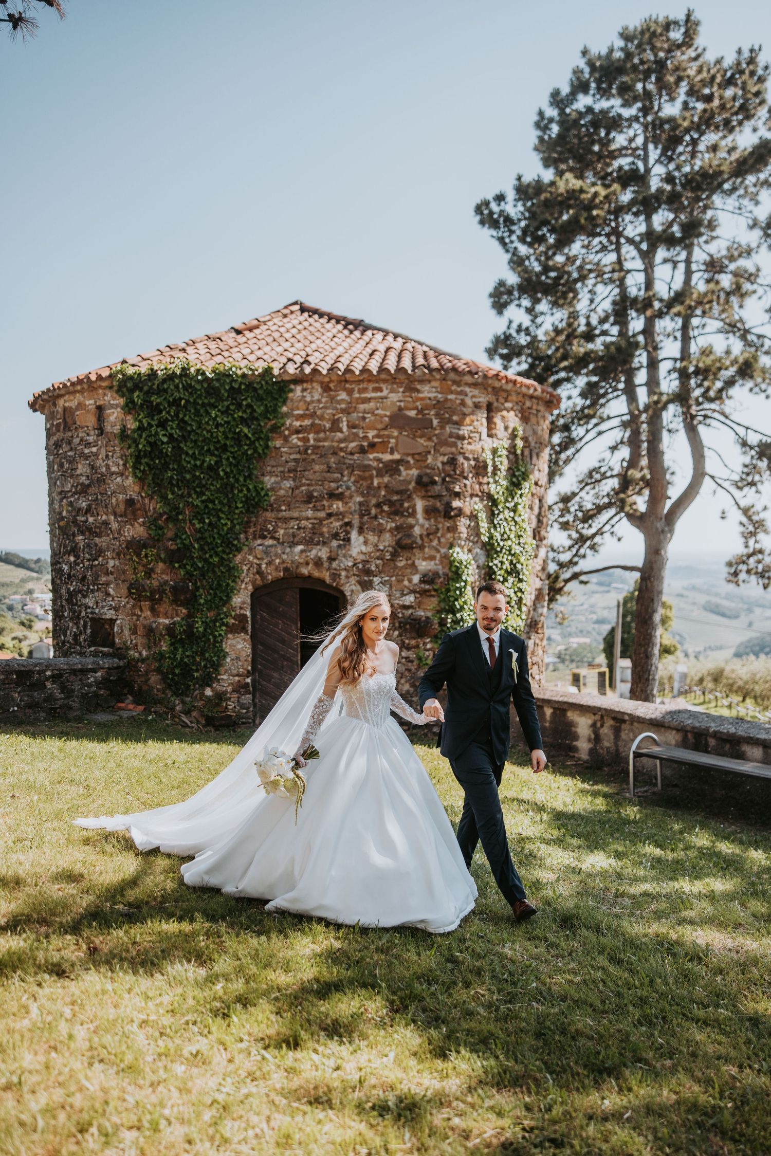 Newlywed couple walks hand in hand on green lawn beside rustic ivy-covered stone chapel on sunny day.