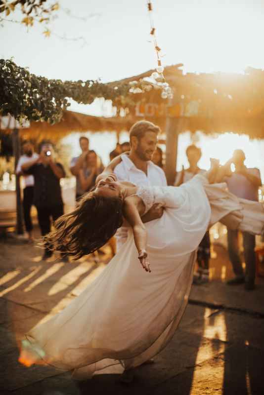 Groom spinning bride in flowing white dress during golden sunset first dance at outdoor wedding.