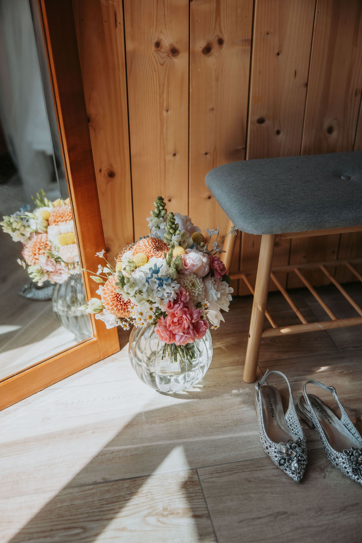 Beautiful wedding bouquet with pink and white flowers in glass vase beside embellished bridal heels on wood floor.