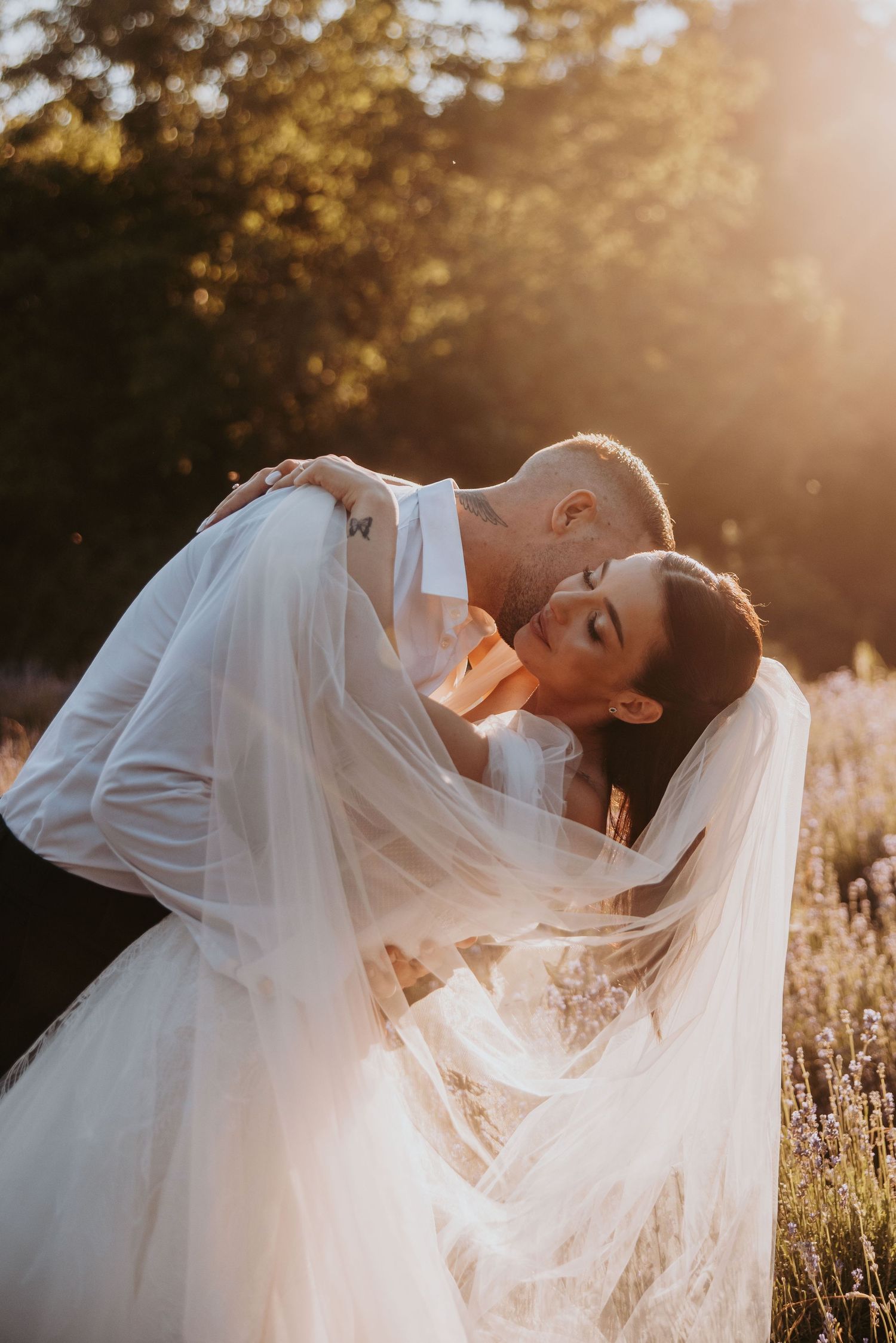 Groom kissing bride in flowing veil during romantic golden sunset embrace in lavender field.