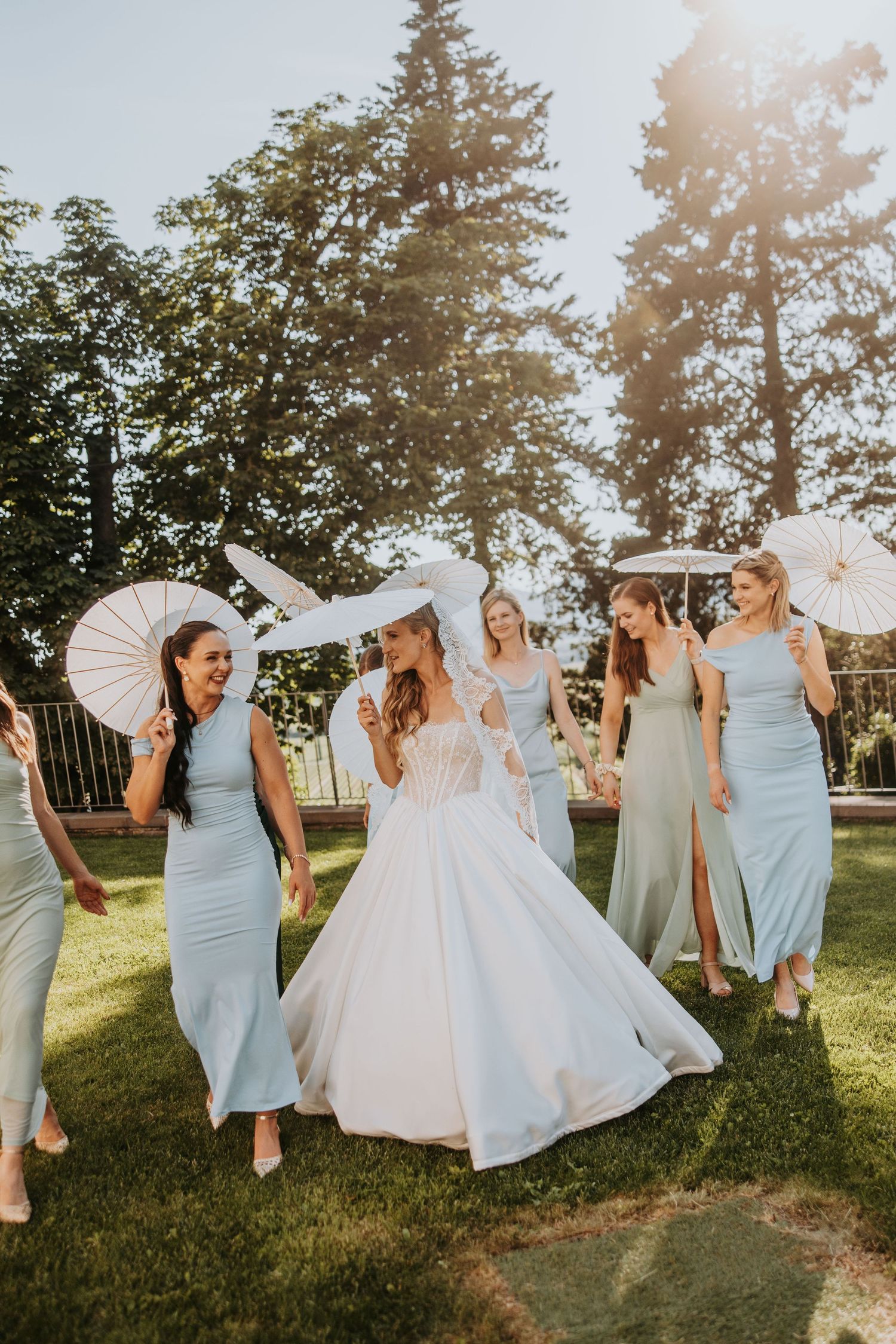 Bride in white ballgown celebrates with bridesmaids in pastel dresses holding white parasols on sunny lawn.