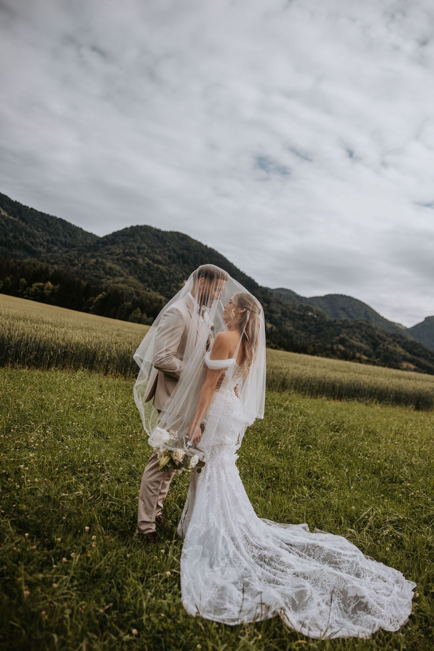 Bride and groom kiss under cathedral veil in lush green field with dramatic mountain landscape behind them.