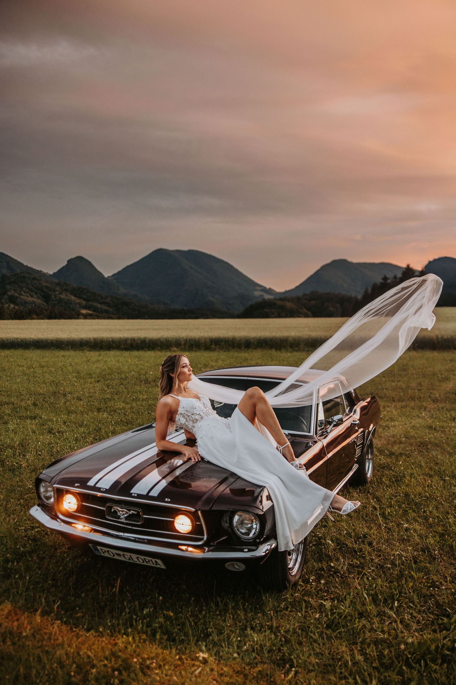 Bride sitting on vintage Ford Mustang hood with veil flowing in wind at golden hour in mountain field.