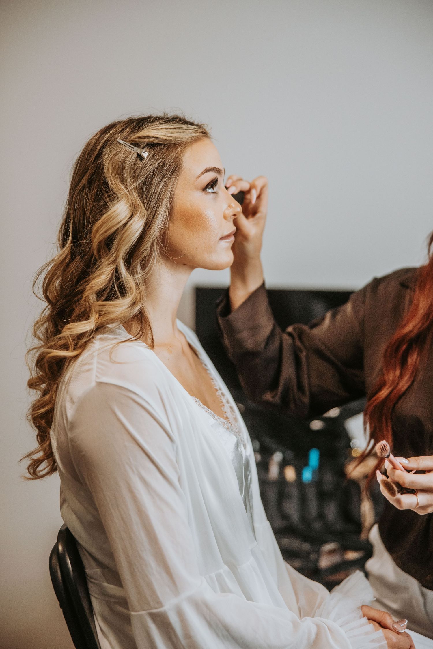 Blonde bride gets professional makeup applied while wearing white robe during wedding preparation.