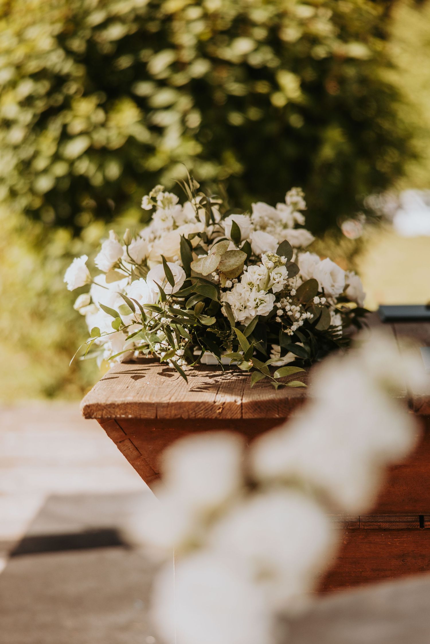 White floral wedding bouquet with greenery resting on a rustic wooden bench outdoors in sunlight.