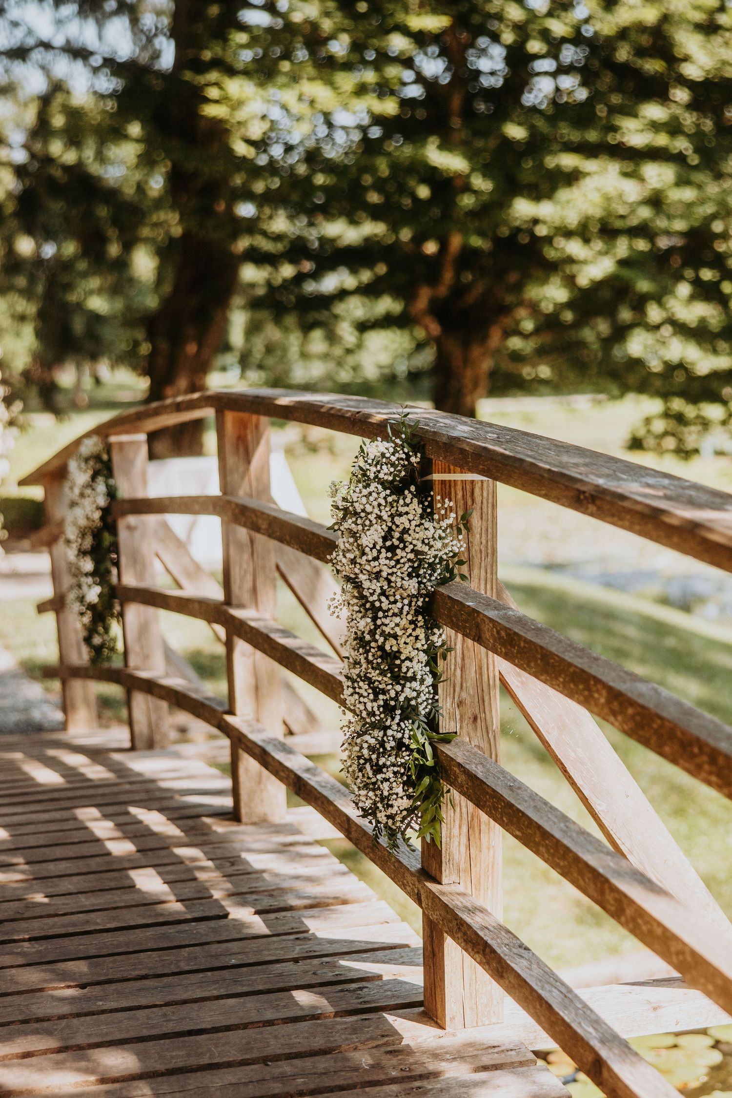 Rustic wooden bridge decorated with white floral garland in a sunlit garden.