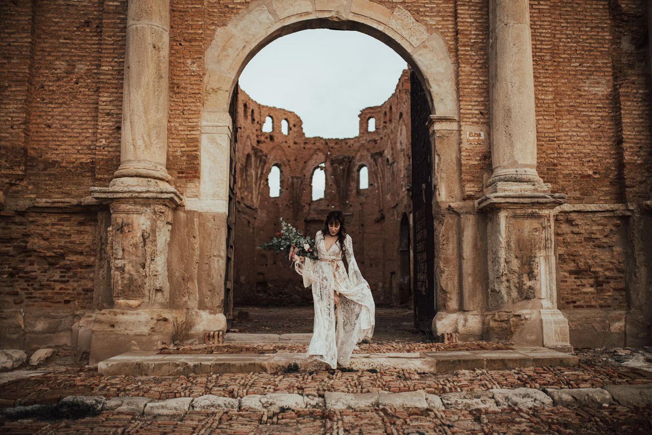 Bride in white lace gown standing in ruins of historic brick archway holding bouquet.