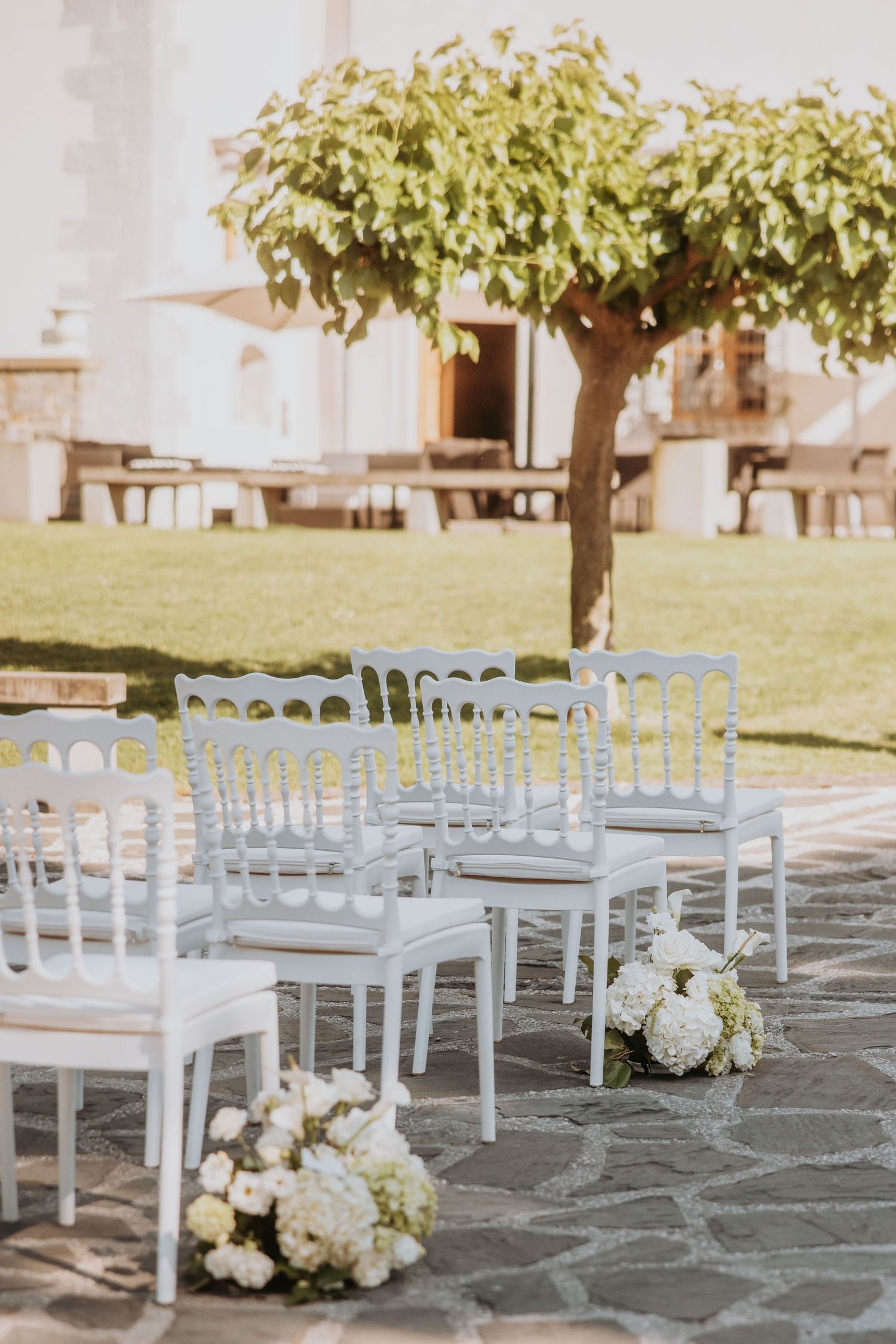 White chairs arranged for an outdoor wedding ceremony on stone patio with white floral arrangements and green tree backdrop.
