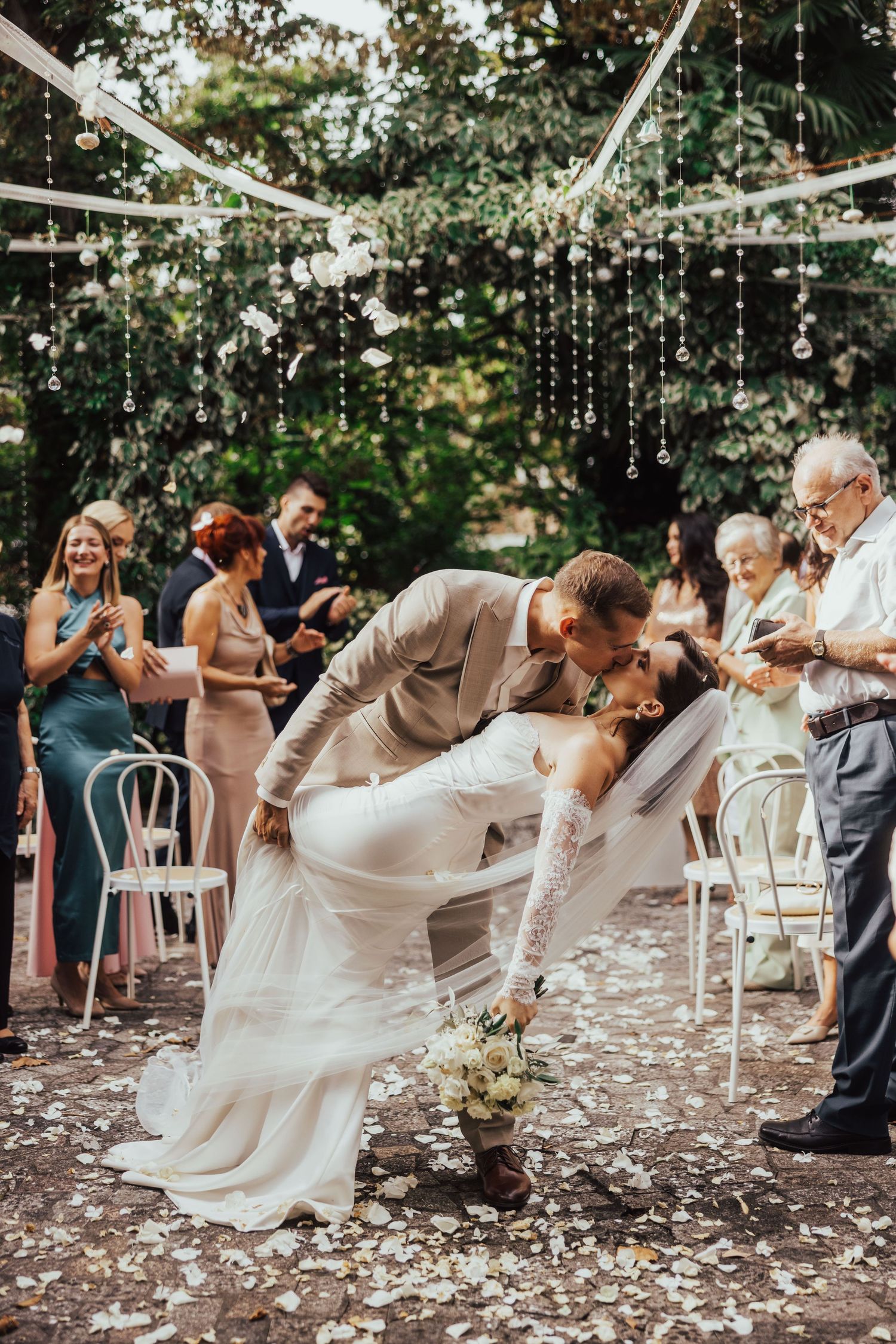 Groom dips bride for romantic kiss at outdoor wedding ceremony as guests celebrate with confetti.