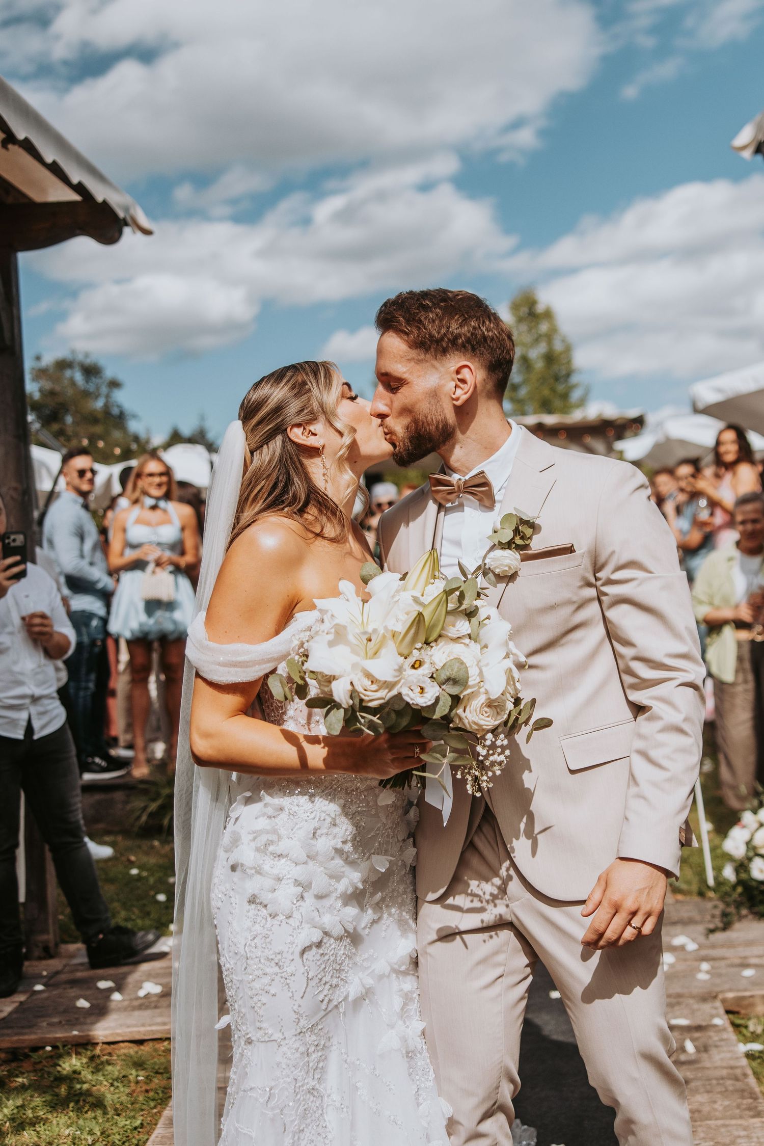 Bride and groom share a kiss holding white floral bouquet at sunny outdoor wedding ceremony.