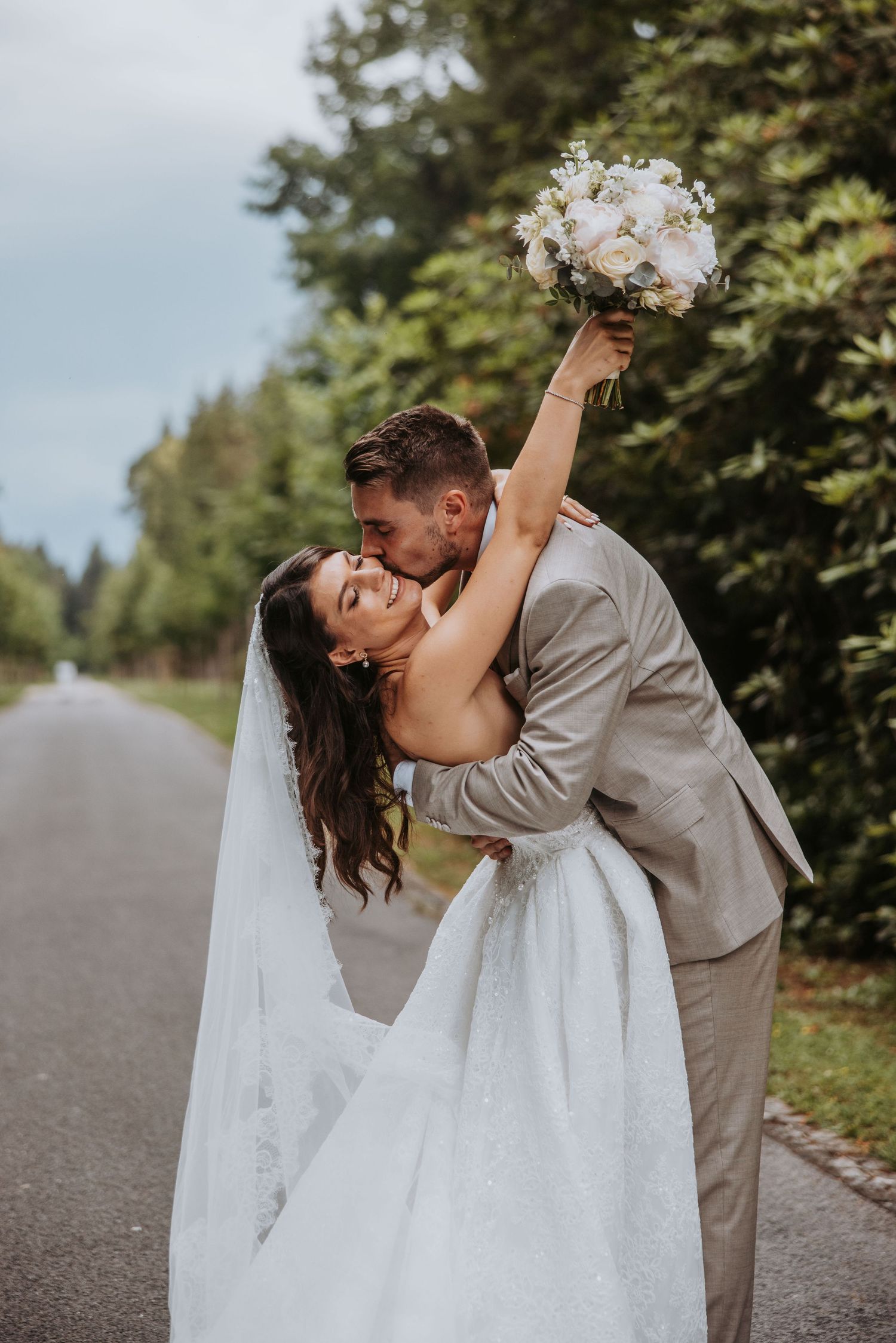 Groom kisses bride while she leans back joyfully holding bouquet aloft on tree-lined road.