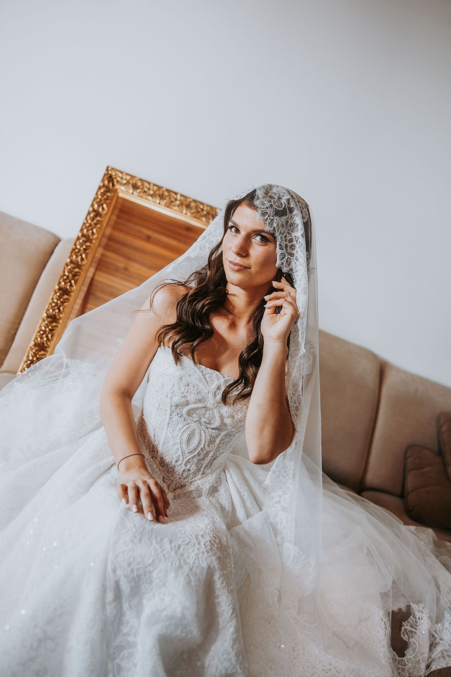 Elegant bride in lace wedding gown and veil sits thoughtfully on a beige sofa.