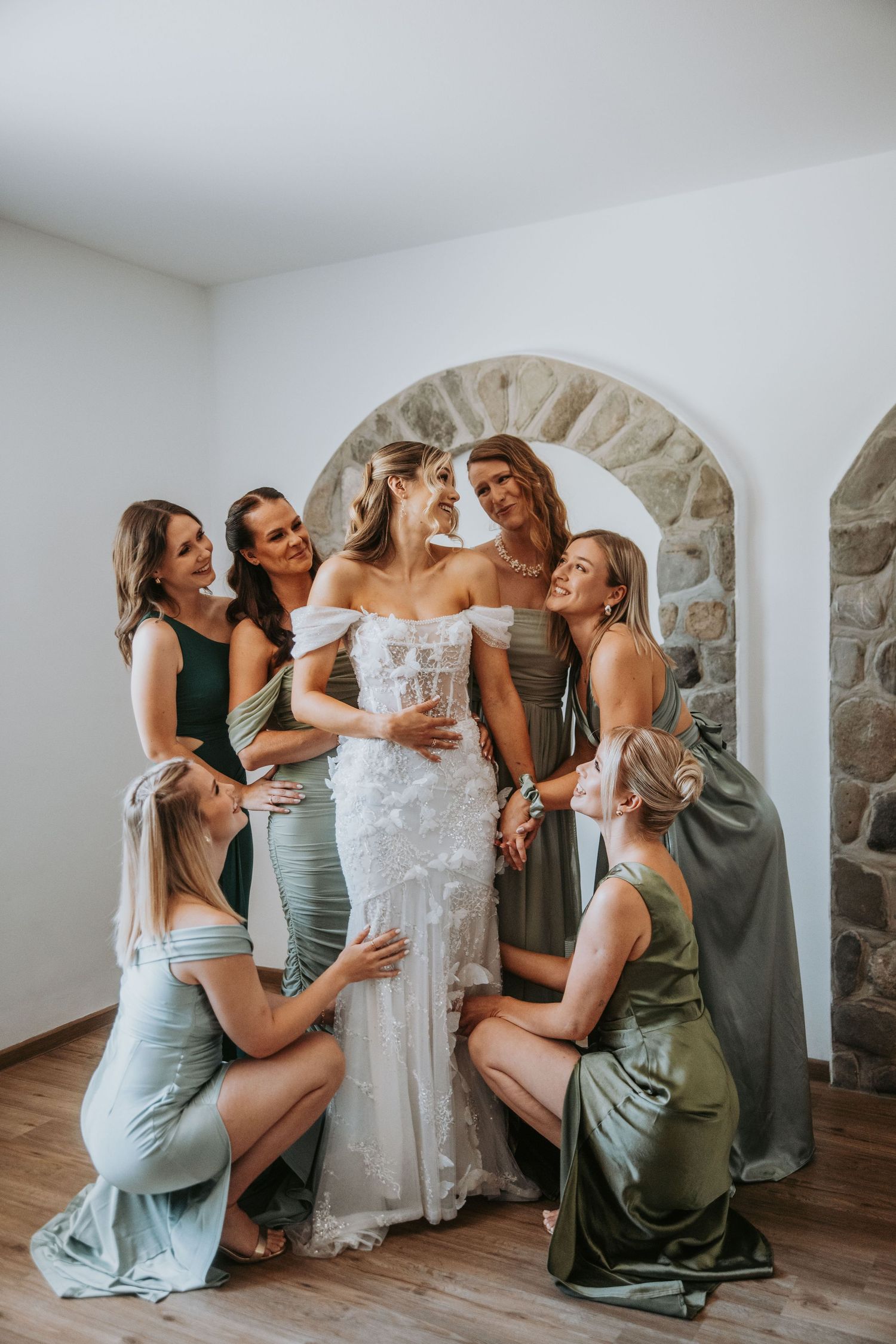Bride in floral lace gown surrounded by bridesmaids in sage green dresses under stone arch.
