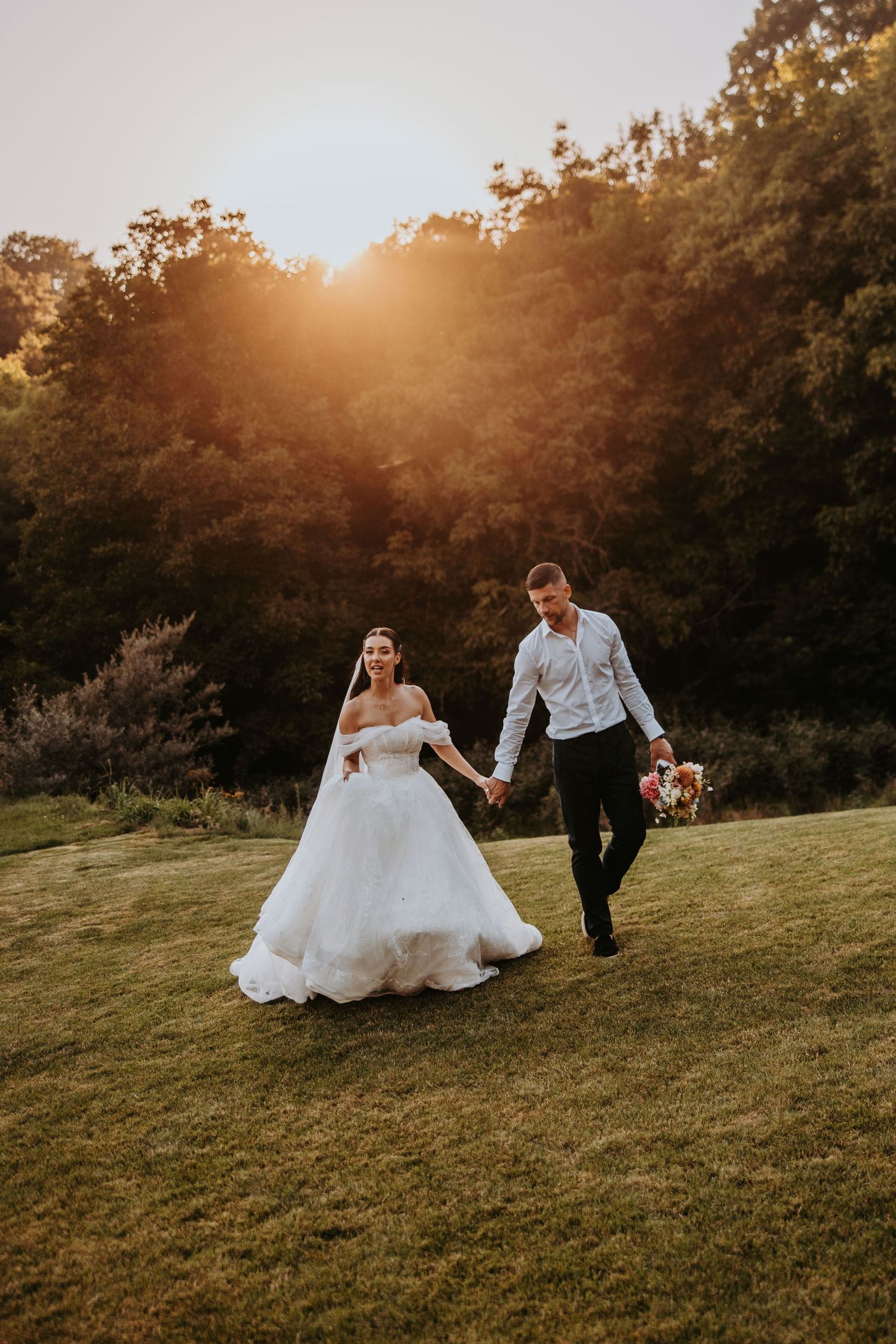 Bride and groom walk hand-in-hand across green lawn at golden sunset with lush trees in background.