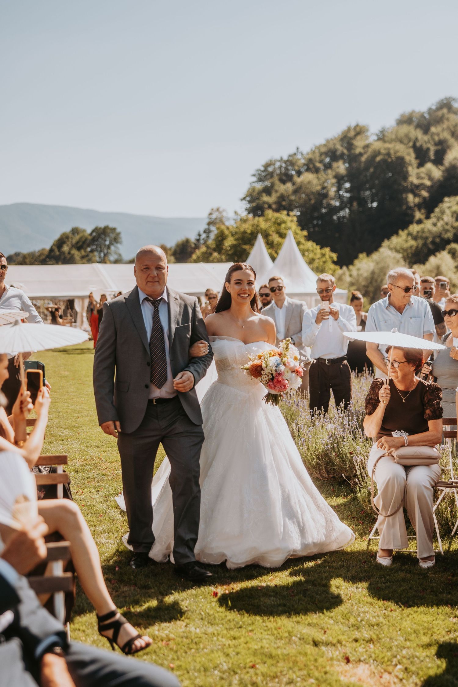 Bride in white gown walks down outdoor aisle with older man, holding colorful bouquet, guests seated alongside.