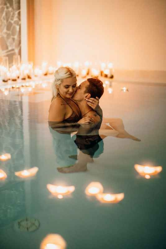 Couple sharing intimate kiss in candlelit pool with floating candles creating romantic ambiance.