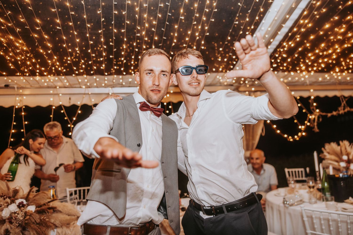 Two men dance and pose energetically under twinkling fairy lights at wedding reception party.