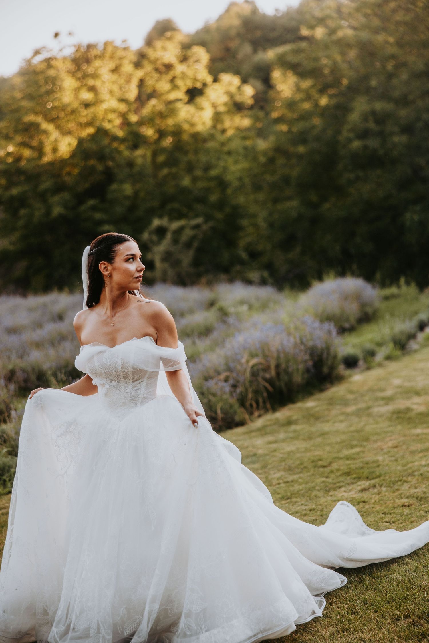 Elegant bride in off-shoulder white ballgown posing in lavender field at golden hour sunset.