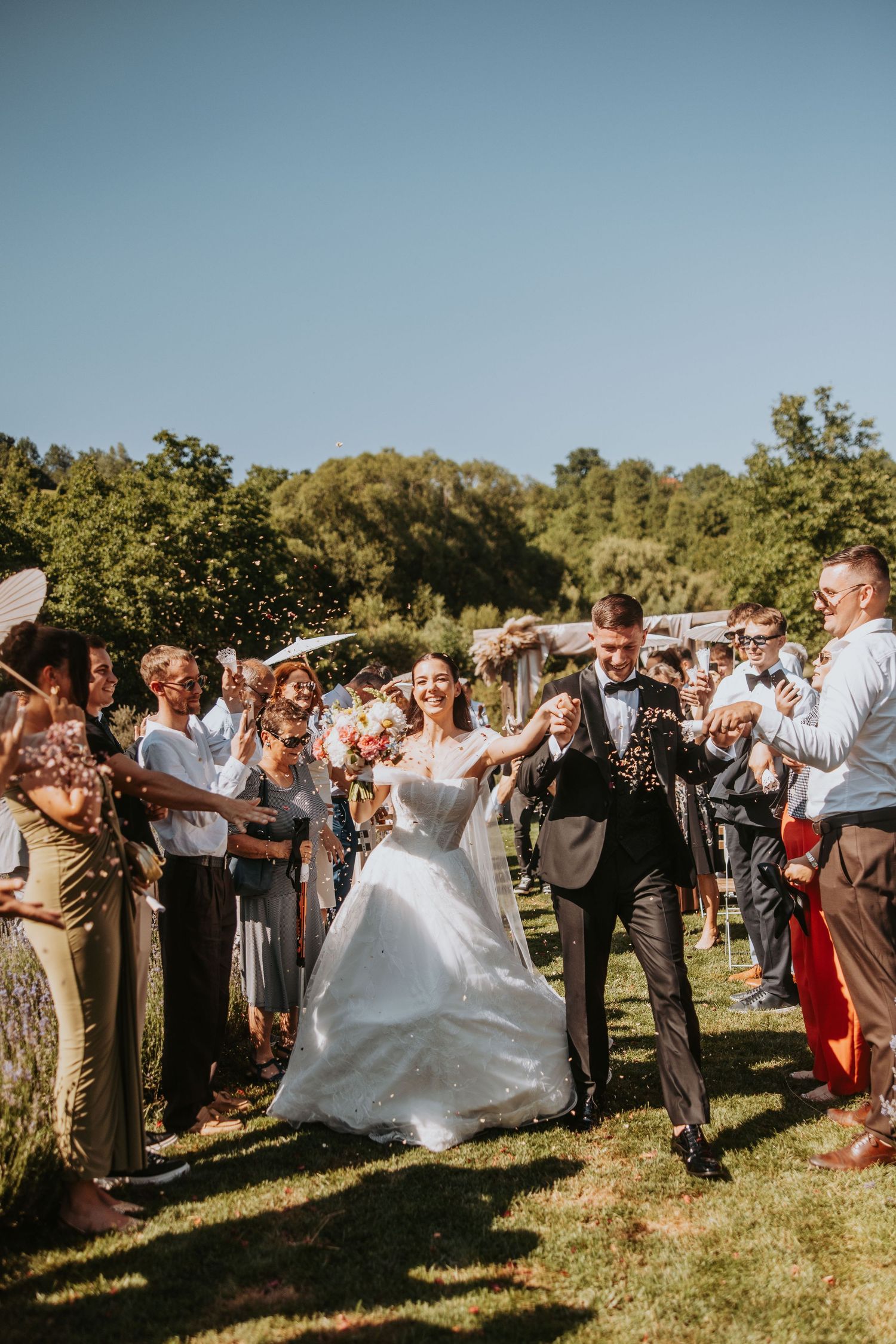 Joyful newlyweds walk through confetti shower surrounded by cheering guests at outdoor wedding.