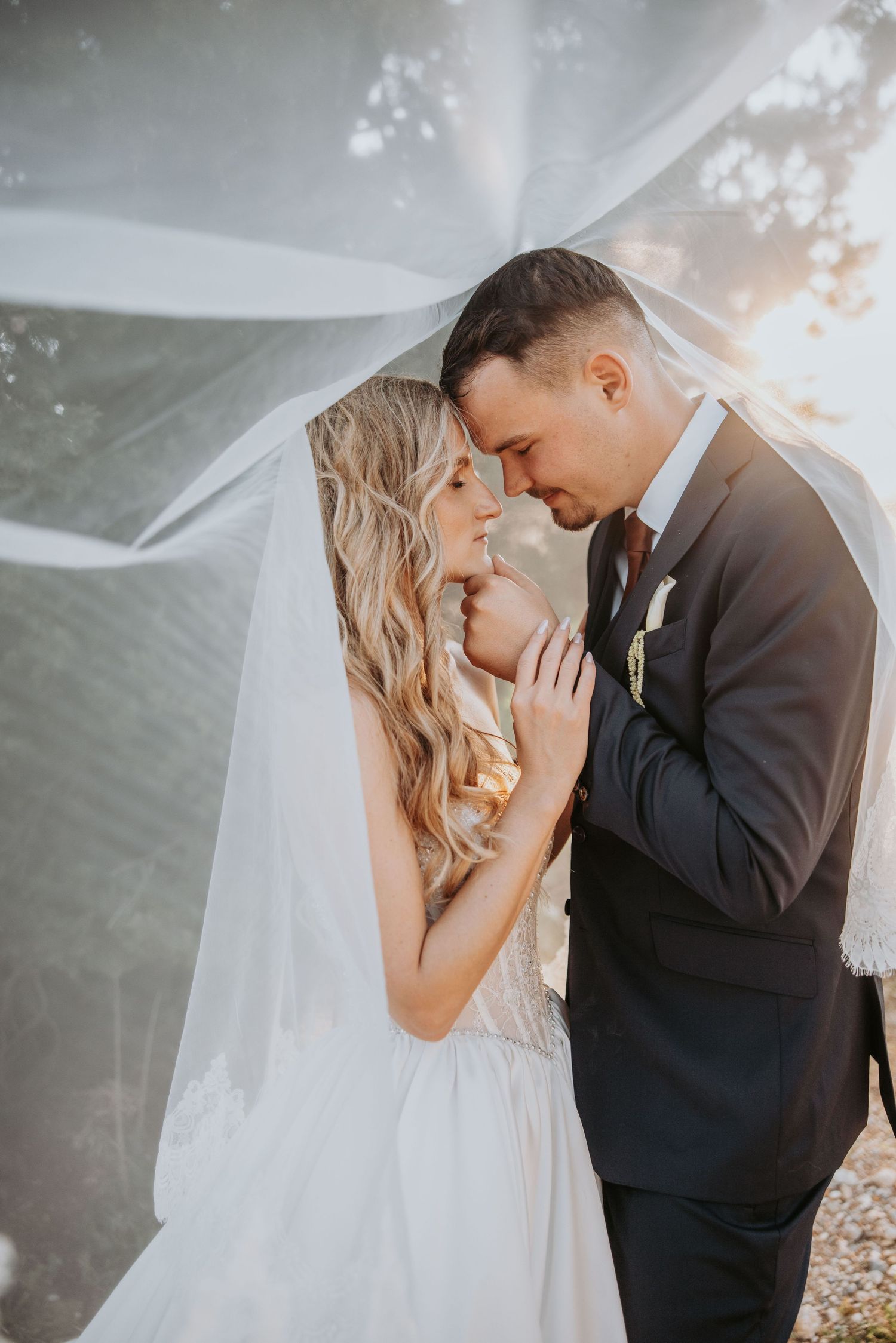Bride and groom standing close under flowing white veil in warm backlit outdoor setting.