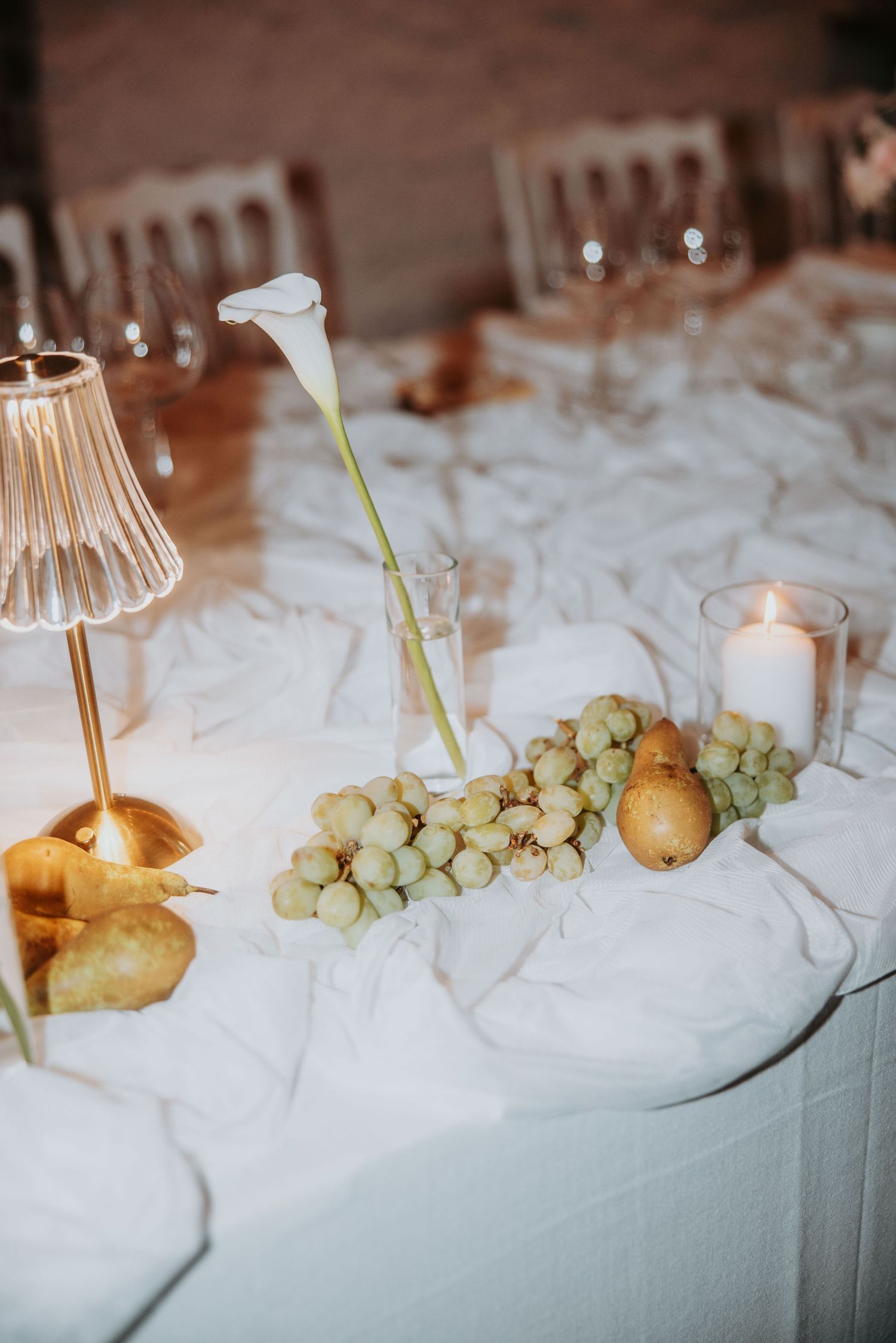 Elegant wedding reception table with white linens, grapes, pears, calla lily, candle and gold lamp.